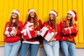 Happy excited group of friends dressed in red sweaters and santa claus hats holding christmas presents while standing near yellow Royalty Free Stock Photo