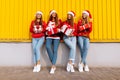 Happy excited group of friends dressed in red sweaters and santa claus hats holding christmas presents while standing near yellow Royalty Free Stock Photo
