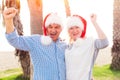 Happy excited mixed race senior couple celebrating xmas on the beach, wearing red Santa`s hats