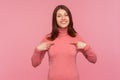 Happy excited woman with brown hair in pink sweater pointing fingers at herself looking at camera with toothy smile, satisfied