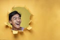 Happy excited Young Asian teenager man poses through torn yellow paper hole, looking at a copy space or blank yellow paper, Royalty Free Stock Photo