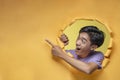 Happy excited Young Asian teenager man poses through torn yellow paper hole pointing a copy space or blank yellow paper, wearing Royalty Free Stock Photo