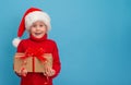 Happy little girl in red sweater and Santa hat holding Christmas gift box on blue background Royalty Free Stock Photo