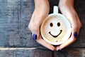 Happy face on cappuccino foam, woman hands holding one cappuccino cup on wooden table