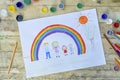 Happy family concept. Drawing on wooden table: father, mother, boy and girl hold hands against background of rainbow and sunny sky