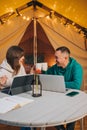 Happy family couple freelancers working laptop on a cozy glamping tent in summer evening. Luxury camping tent for outdoor holiday