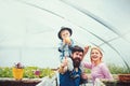 Happy family in greenhouse. Father in blue vest holding his son on shoulders. Cute kid in hat eating apple while his mom