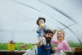 Happy family in greenhouse. Father in blue vest holding his son on shoulders. Cute kid in hat eating apple while his mom