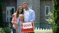 Happy family holding sold sign, standing against new bought house, thumbs up