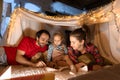 Happy family, mother, father and daughter lying inside self-made hut, tent in room in the evening and reading book Royalty Free Stock Photo