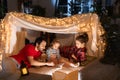 Happy family, mother, father and daughter lying inside self-made hut, tent in room in the evening and reading book Royalty Free Stock Photo