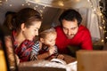 Happy family, mother, father and daughter in pajamas lying inside self-made hut, tent in room in the evening and reading Royalty Free Stock Photo