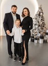 Happy family near fir-tree. Mom in a long black dress, dad in a suit and son in a white shirt, hugging and smiling Royalty Free Stock Photo