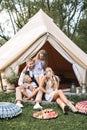 Happy family on picnic, sitting on green grass near the big white tipi tent in forest or park. Father, mother and two Royalty Free Stock Photo