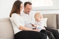 Happy family of three is enjoying at home. Parents sitting on the sofa and looking at their beautiful baby girl playing with cell Royalty Free Stock Photo