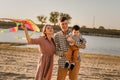 Happy family walking on sandy beach of river. Father, mother holding baby son on hands and playing with kite Royalty Free Stock Photo