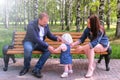 Baby girl trying to make first steps from mom to dad sitting on bench in park. Royalty Free Stock Photo