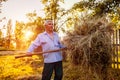 Happy farmer man gathers hay with pitchfork at sunset in countryside