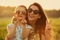 Happy fashion kid girl embracing her mother in trendy sunglasses and showing victory sign by two fingers on nature background. Royalty Free Stock Photo