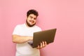 Happy fat man in white t-shirt uses a laptop with a smile on his face on a pink background, looks into the screen. Charismatic Royalty Free Stock Photo