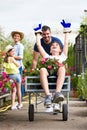 Happy father and his son playing with a wheelbarrow while girl and her mother looking them in the greenhouse. Royalty Free Stock Photo