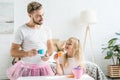 Happy father in pink tutu skirt looking at cute little daughter pretending to have tea party