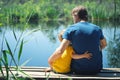 Happy father and son hugging sitting on wooden pier near lake on sunny summer day. Dad and child boy spend time together on wharf Royalty Free Stock Photo