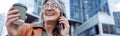 Happy female pensioner with cup of drink n talks on mobile phone on modern city street Royalty Free Stock Photo