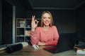 Happy female student sitting at home at desk with laptop and books and posing at camera with smile on face and showing ok gesture Royalty Free Stock Photo