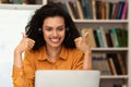Happy female teacher giving lesson on web using laptop and gesturing thumbs up at webcamera, sitting in library Royalty Free Stock Photo