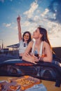 Happy females having fun and drinking soda in glass bottles, posing in yellow car with french fries and pizza on its