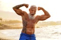 Happy fit senior man showing his muscle on the beach after outdoor workout, during sunset time