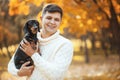 Happy free time with beloved dog! Handsome young man staying in autumn park smiling and holding cute puppy dachshund.