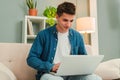 Happy freelancer man working at home with a laptop on a sofa at daytime. Young guy smiling and browsing on internet Royalty Free Stock Photo