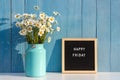 Happy Friday words on black letter board and bouquet of chamomile flowers on table against blue wooden wall. Concept Hello Friday