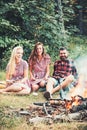 Happy friends at campfire. Bearded man and women smile at bonfire. Hipster in palid shirt and girls in vintage dresses Royalty Free Stock Photo