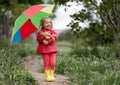 A girl of three years old in a pink jacket, rubber boots and with a multi-colored umbrella walks alone in a green spring park in t Royalty Free Stock Photo