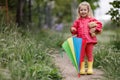 A girl of three years old in a pink jacket, rubber boots and with a multi-colored umbrella walks alone in a green spring park in t Royalty Free Stock Photo