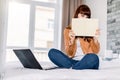 Happy funny beautiful woman in jeans and yellow casual shirt, hiding her face with notepad, while working on a laptop Royalty Free Stock Photo