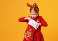 Happy funny child girl in red Christmas reindeer costume with gift surprise sock on yellow   background Royalty Free Stock Photo