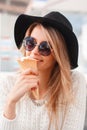 Happy funny hipster young woman in elegant black hat in a knitted sweater sits and eats sweet ice cream in a summer cafe.