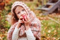 Happy funny kid girl in cozy knitted scarf eating fresh apple in autumn garden