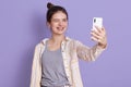 Happy funny woman posing isolated over lilac background, winks eye and taking selfie, woman with dark hair and bun standing Royalty Free Stock Photo