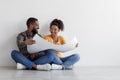 Happy funny young african american guy and lady look at blueprints at home, sit on floor over white wall background