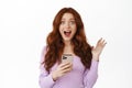 Happy ginger girl screams, holds smartphone and smiling, celebrating big news, online promotion, standing in blouse