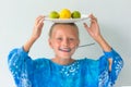 Happy girl with blue eyes in a blue dress holding a spoon in his mouth, a plate with lime and lemon on her head on a white