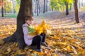 Happy girl with a bouquet of maple yellow autumn leaves sitting under a tree in a sunny city park. Life style Royalty Free Stock Photo