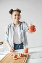 Happy girl with buns smiling stretching glass with grapefruit detox smoothie to somebody. White wall background. Healthy