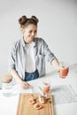 Happy girl with buns smiling stretching glass with grapefruit detox smoothie to somebody. White wall background. Healthy