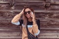 Happy girl calls by phone, background wooden building fence, in beige sweater and with bag over his shoulder. Sunglasses Royalty Free Stock Photo